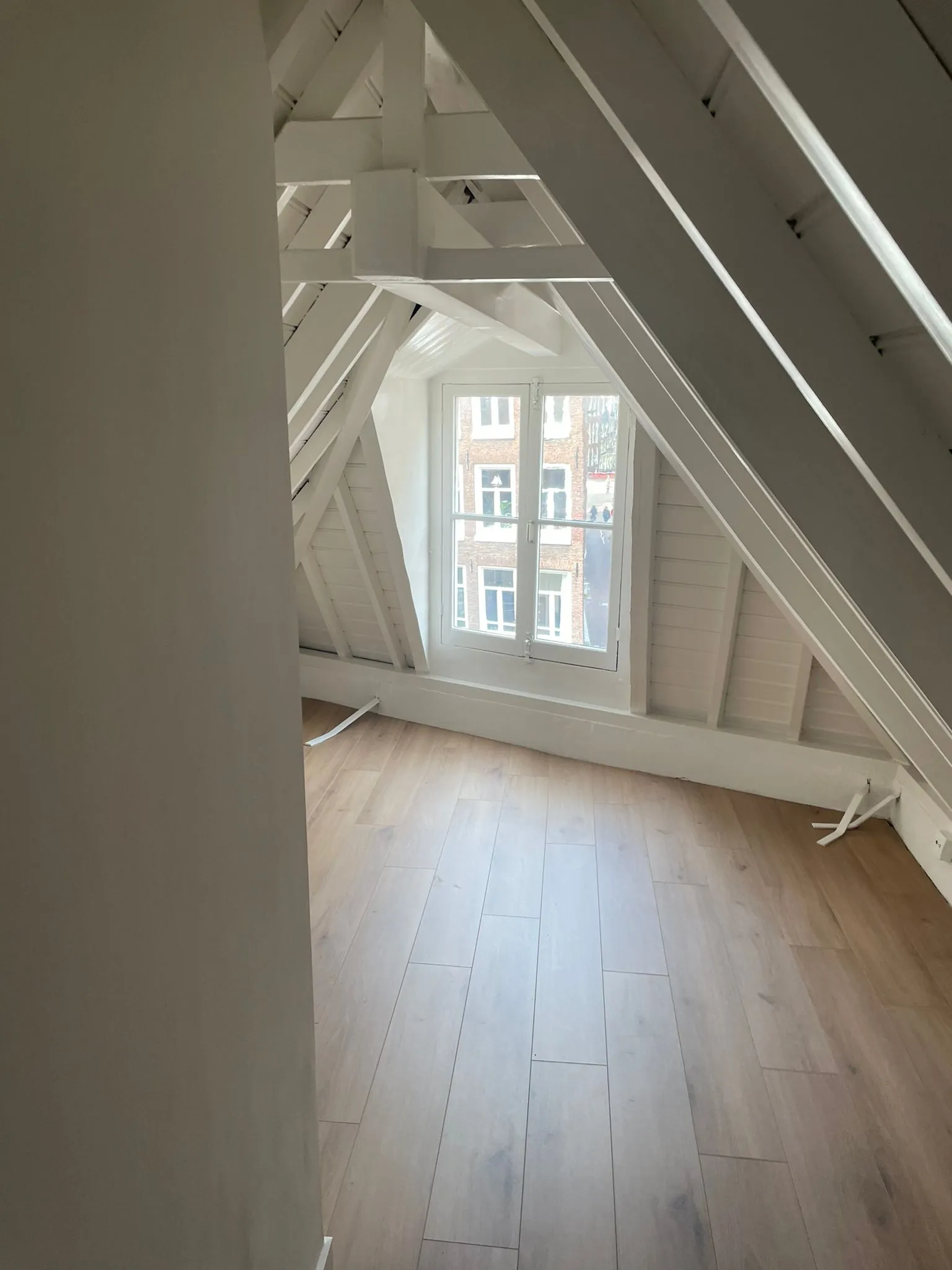 Zolderkamer met houten vloer, witte balken en een raam met uitzicht op bakstenen woningen aan de Korte Kolksteeg.